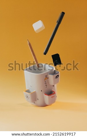 Ceramic design pencil holder with ballpoint pen, eraser, memory card and pencil flying on yellow background. Vertical.