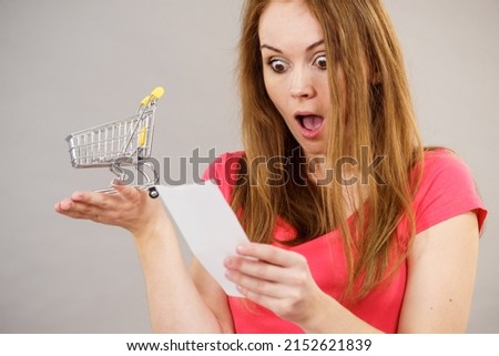 Shocked woman holding list of products or bill and small shopping cart bag trolley. Female in shop buying groceries. High cost of living.