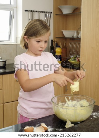 Girl (6-8) standing in kitchen, dipping finger into bowl of cake mix, holding spoon