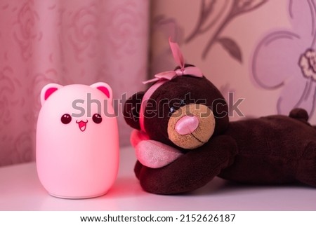 The baby cute pink bear-shaped night lamp with eyes and ears near a toy teddy bear with a bow on its head and a heart in its hands on the bedside table in the baby girl room glowing in the dark