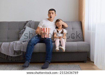Portrait of young adult man watching horror movie at home with his daughter, covering eyes to his kid, sitting on sofa and holding popcorn in bucket, expressing shock, being very impressed.
