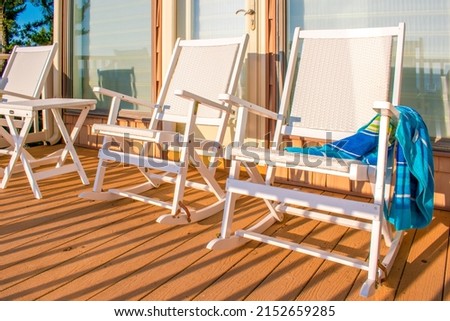 Rocking chairs on a wooden porch at a beach house with a towel draped over the arm of one chair in a bold and bright realistic style. Royalty-Free Stock Photo #2152659285