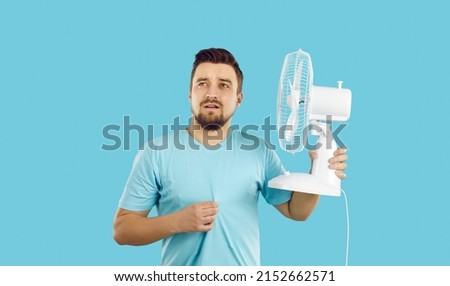 Man suffers from summer heat at home. Guy with broken air conditioner in his house using bad electric fan and sweating. Man in T shirt feeling hot and holding fan with sad face expression, studio shot Royalty-Free Stock Photo #2152662571