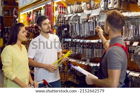 Family in hardware store. Seller advises couple of customers on type of spanners in building materials store. Husband and wife buy tools for repairs, listen carefully to employee of store in overalls. Royalty-Free Stock Photo #2152662579