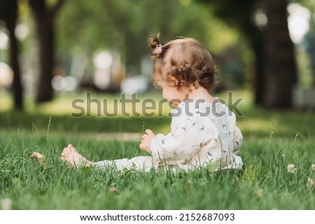 cute little smiling girl with a funny hairstyle is sitting on a blooming green lawn in the park. child plays and walks outdoors. lifestyle. space for text. High quality photo