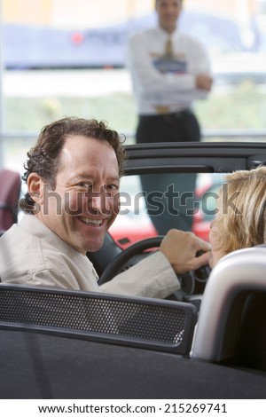 Couple sitting in new convertible in car showroom, salesman in background, man smiling, portrait