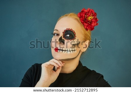 Beautiful redhead woman with frightening make-up in a black dress with a red flower in her hair on a green background. Day of the Dead in Mexico.