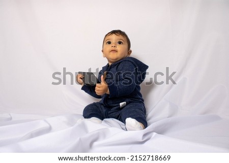 Baby with a cell phone in one hand and making a positive sign with the other on a white cloth in mention of understanding the technologies. Children and communication through mobile devices.