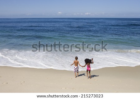 Boy and girl (5-7) holding hands on sandy beach near water's edge, Atlantic Ocean in background, rear view