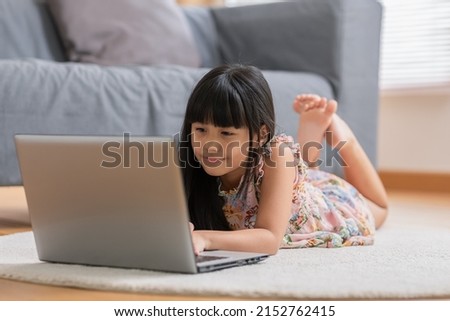 Cute little asian girl uses laptop while prone at the sofa in the living room. Child surfing the internet on computer, browses through internet and watches cartoons online at home