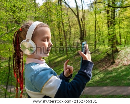 Adorable child girl in headphones learning language listening to music using smartphone outdoor sunny day. Blogger kid with gadget distance school online lesson chat cell phone conference in the park.