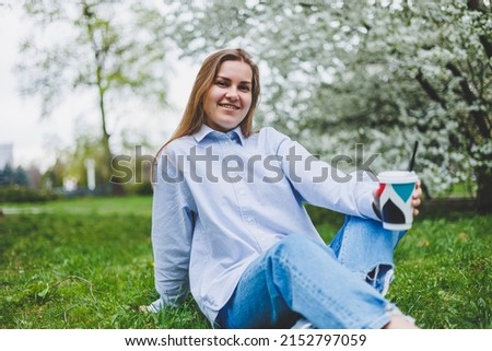 Young hipster woman in denim jacket is resting on green lawn in park, drinking coffee, relaxing outdoors. Lifestyle, coffee break. Selective focus on female hands holding disposable cup of coffee.