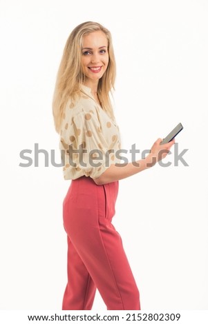 cute Caucasian blue-eyed blonde girl with a pretty smile wearing a shirt and pink trousers standing sideways and holding a phone studio shot white background medium full shot copy space. High quality