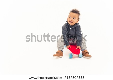 cute curly little boy sitting on the red skateboard and looking up excited studio shot full shot white background copy space. High quality photo