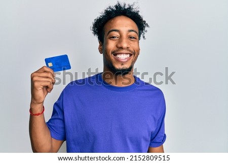 Young african american man with beard holding credit card looking positive and happy standing and smiling with a confident smile showing teeth 