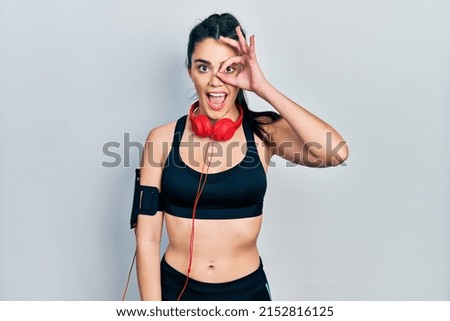 Young hispanic girl wearing gym clothes and using headphones doing ok gesture with hand smiling, eye looking through fingers with happy face. 