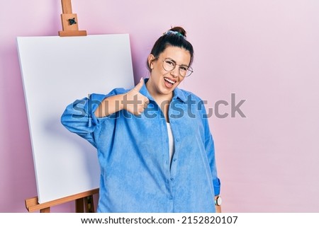 Young hispanic woman standing by painter easel stand doing happy thumbs up gesture with hand. approving expression looking at the camera showing success. 