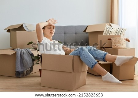 Indoor shot of tired dark haired woman wearing white t shirt and jeans sitting in a cardboard box, relocating to a new house, being exhausted to unpack personal piles. Royalty-Free Stock Photo #2152823653