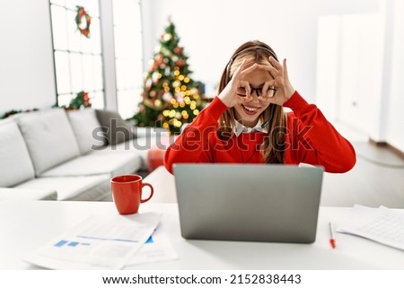 Young caucasian girl sitting on the table working using laptop by christmas tree doing ok gesture like binoculars sticking tongue out, eyes looking through fingers. crazy expression. 