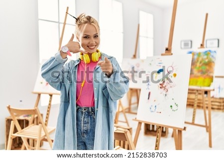 Young caucasian girl at art studio smiling doing talking on the telephone gesture and pointing to you. call me. 