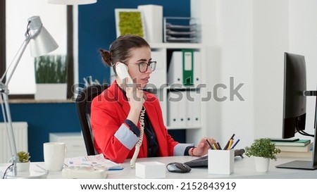 Company secretary having remote conversation on landline phone, working on financial development strategy with computer. Female employee answering office telephone call for startup work. Royalty-Free Stock Photo #2152843919