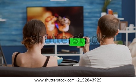 Couple at home watching online tutorials on green screen smartphone while enjoying free time sitting on couch. Man and woman looking at webinar or social media videos on chroma key mobile phone.