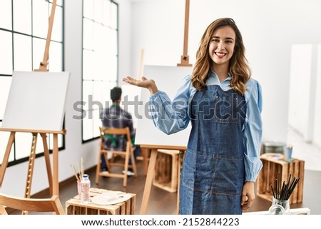 Young artist woman at art studio smiling cheerful presenting and pointing with palm of hand looking at the camera. 