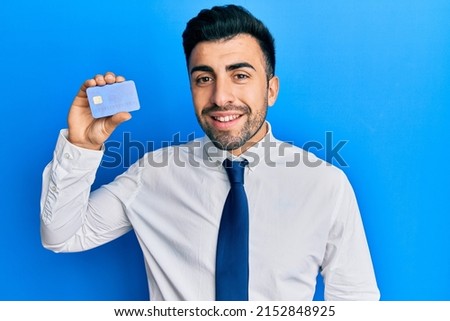 Young hispanic man wearing business clothes holding credit card looking positive and happy standing and smiling with a confident smile showing teeth 