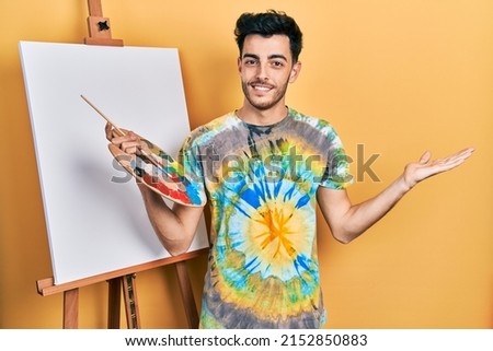 Young hispanic man standing drawing with palette by painter easel stand smiling cheerful presenting and pointing with palm of hand looking at the camera. 