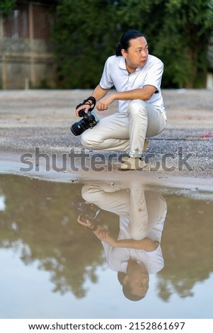 Asian man holds a big black mirrorless camera medium format type on cement asphalt ground in front of water reflex.