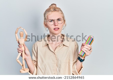 Beautiful caucasian woman with blond hair holding small wooden manikin pencils clueless and confused expression. doubt concept. 