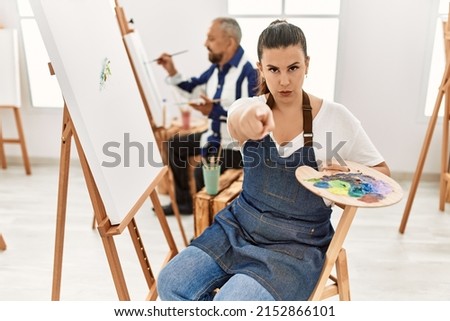 Young artist woman at art studio pointing with finger to the camera and to you, confident gesture looking serious 