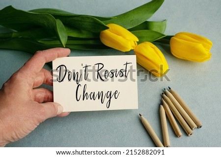 The hand of a young girl close-up holds a card with the inscription Don't resist change on a light blue background along with yellow tulip flowers. View from above. Conceptual image.