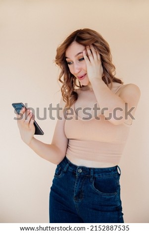 Photo of a young Caucasian girl with blue eyes using her mobile phone with a serious face to make a call. A woman is talking on the phone against a beige wall. Empty space for the text.