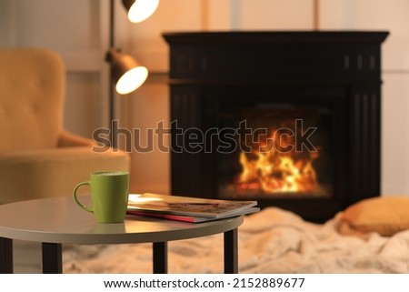 Cup of hot drink and magazines on table near fireplace at home, space for text. Cozy atmosphere