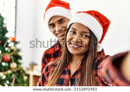 Young latin couple wearing christmas hat making selfie by the smartphone at home.