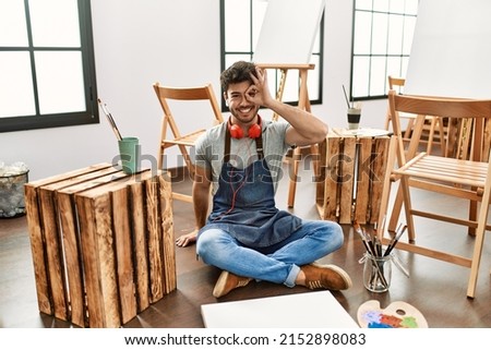 Young hispanic man sitting at art studio doing ok gesture with hand smiling, eye looking through fingers with happy face. 
