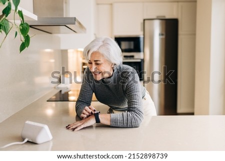 Senior woman smiling happily while using smart devices in her kitchen. Cheerful elderly woman using a home assistant to perform tasks at home. Royalty-Free Stock Photo #2152898739