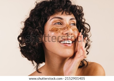 Confident young woman looking away with a smile while applying coffee scrub on her face. Happy young woman exfoliating her facial skin using a cleansing coffee mask. Royalty-Free Stock Photo #2152901405
