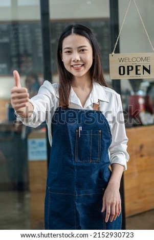 A young Asian businesswoman who owns an apron coffee shop stands in front of a shop with a sign that reads open.