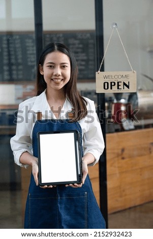 An Asian businesswoman who owns a coffee shop holds a tablet with a white screen and an apron standing in front of her shop with a sign that reads open.