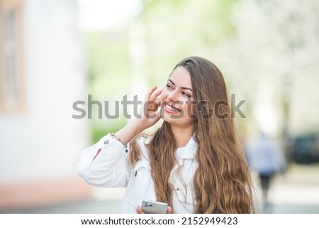 A young beautiful woman with long hair in comfortable light casual clothes listens to music using bluetooth headphones, walks in the center of a European city. A cute model is shopping.