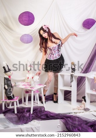 The girl, like a doll Alice in Wonderland, pours tea for dolls to rabbits from the tea set. Purple shades, white background.