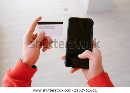 Point of view cropped close up view photo of man hands wearing red hoodie using blank credit card and smart telephone. Online payment concept.