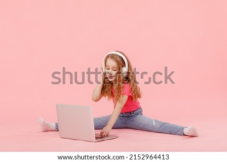 adorable child girl is sitting in the twine next to a laptop. kid in white headphones stretching and learns homework or watching cartoons, listening music.