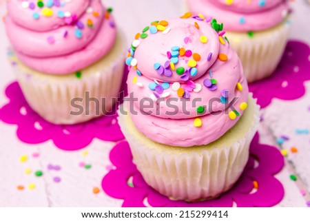 3 fresh baked vanilla cupcakes with pink swirled strawberry frosting topped with colorful sprinkles sitting on flower cut outs