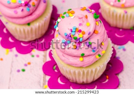 3 fresh baked vanilla cupcakes with pink swirled strawberry frosting topped with colorful sprinkles sitting on flower cut outs