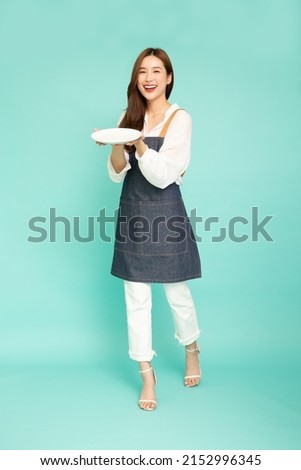 Young Asian woman housewife wearing kitchen apron cooking and holding empty white plate or dish isolated on green background, Full length composition Royalty-Free Stock Photo #2152996345