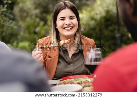 chubby girl smiles and chats with multiethnic group of friends eating meat skewers in the backyard - A chubby woman sits at the picnic table on a cool summer day eating grilled food