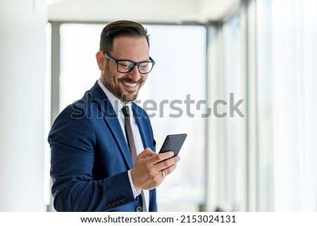 Handsome businessman wearing eyeglasses using smart phone and smiling while standing near the office window, Copy Space Royalty-Free Stock Photo #2153024131